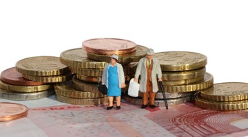 Deux petites figurines, un homme et une femme, se tiennent côte à côte sur une pile de pièces et de billets en euros. La femme porte une robe bleue et tient un sac, tandis que l'homme porte un chapeau et tient une canne.