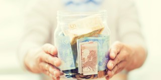 Un bocal en verre rempli de billets et de pièces de monnaie, tenu par une personne avec les mains tendues.