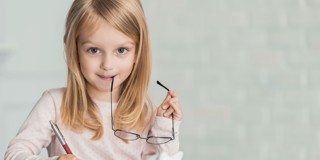 Un enfant aux longs cheveux blonds tient un crayon et porte des lunettes, tout en écrivant à un bureau sur lequel se trouve une tirelire.