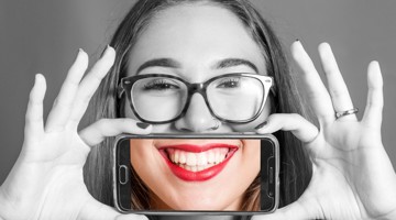 Une femme tient un smartphone devant son visage avec les deux mains, tout en regardant la caméra.