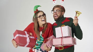 Deux personnes vêtues de façon festive, l'une portant un chapeau d'elfe et l'autre ayant une cloche, se tiennent ensemble près de plusieurs cadeaux emballés.