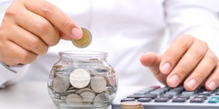 Une main place une pièce dans un bocal en verre plein de pièces, tandis que l'autre main utilise une calculatrice.