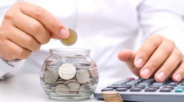 Une main place une pièce dans un bocal en verre plein de pièces, tandis que l'autre main utilise une calculatrice.