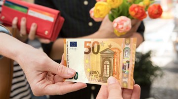 Une main tend un billet de cinquante euros à une autre main, tandis qu'à l'arrière-plan, une personne se tient avec un bouquet de fleurs.