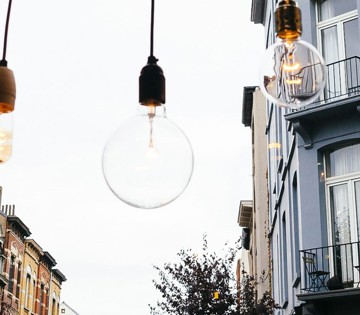 Des lampes suspendues au design varié sont accrochées au-dessus d'une rue bordée de bâtiments et d'arbres en arrière-plan.
