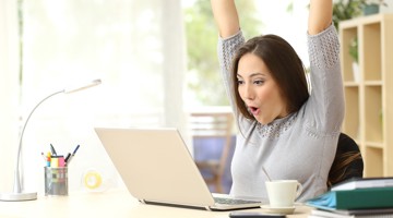 Een persoon met opgestoken armen zit achter een laptop aan een bureau, met een kopje koffie en schrijfmaterialen in de buurt.