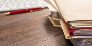 Des piles de classeurs sur un bureau en bois, avec un clavier et un stylo derrière.