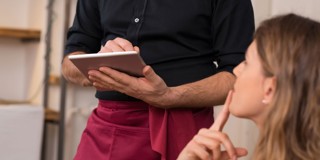 Un serveur en chemise noire avec un tablier bordeaux prend une commande sur une tablette, tandis qu'un client écoute attentivement.