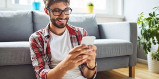 Man in een geruit overhemd zit op de vloer met een smartphone in zijn handen, omringd door een lichte woonkamer met planten en een bank.