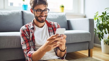 Man in een geruit overhemd zit op de vloer met een smartphone in zijn handen, omringd door een lichte woonkamer met planten en een bank.
