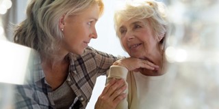 Een jongere vrouw leunt naar een oudere vrouw terwijl ze dicht bij elkaar zitten, en lijkt rustig iets te vertellen met een empathische en aandachtige uitdrukking.