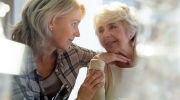 Een jongere vrouw leunt naar een oudere vrouw terwijl ze dicht bij elkaar zitten, en lijkt rustig iets te vertellen met een empathische en aandachtige uitdrukking.