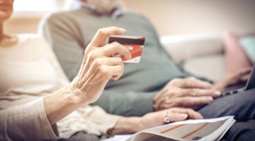 Une femme âgée tient une carte de crédit en étant assise sur un banc à côté d'un homme âgé, avec un ordinateur portable et un magazine devant elle.