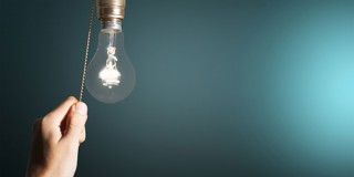 Een hand trekt aan het koord van een gloeilamp die tegen een lichte achtergrond hangt.