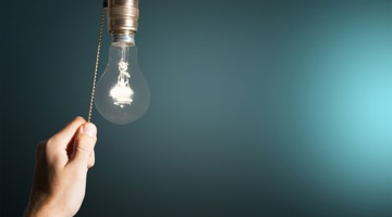 Een hand trekt aan het koord van een gloeilamp die tegen een lichte achtergrond hangt.