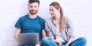 Twee mensen zitten tegen een muur, met een laptop op schoot en een smartphone in de hand, terwijl ze beiden naar het laptopscherm kijken.