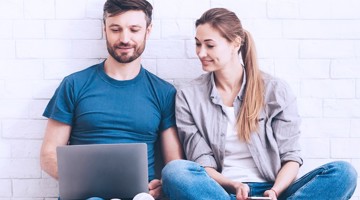 Twee mensen zitten tegen een muur, met een laptop op schoot en een smartphone in de hand, terwijl ze beiden naar het laptopscherm kijken.