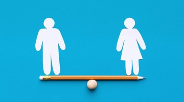 Une poutre d'équilibre avec une boule en bois motivée au centre, sur laquelle repose un crayon. Sur le côté gauche, un symbole en papier représentant un homme, et sur le côté droit, un symbole en papier représentant une femme.