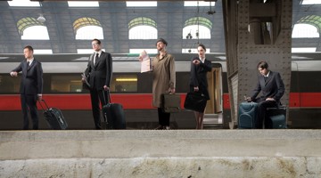Vijf mensen in formele kleding staan met koffers op een treinstation, terwijl er een trein op de achtergrond voorbijrijdt.