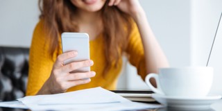 Vrouw in een gele trui kijkt naar haar smartphone, terwijl er een kop koffie en een stapel papieren op de tafel staan.