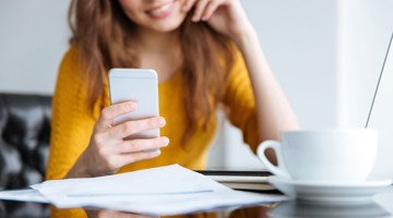 Vrouw in een gele trui kijkt naar haar smartphone, terwijl er een kop koffie en een stapel papieren op de tafel staan.