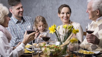 Een gezin zit aan tafel en geniet van een maaltijd met diverse gerechten, terwijl ze glazen rode wijn vasthouden en vrolijk met elkaar praten. Voor hen staan borden met eten en een vaas met gele narcissen.