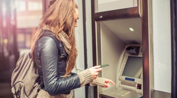 Vrouw met lange haren die een bankpas in een geldautomaat steekt, terwijl ze een rugzak op haar schouder heeft.