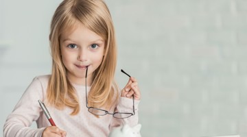 Een kind houdt een pen en een bril vast en kijkt met een neutrale achtergrond recht in de camera.