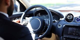 Man in een donker pak die een auto bestuurt, met focus op het stuur en dashboard.
