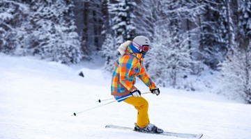 Skiër in een kleurrijke winteroutfit glijdt over een besneeuwde helling, omringd door bomen.
