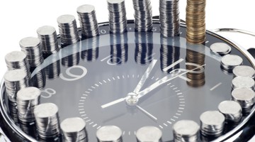 Een klok met een zwarte wijzerplaat, omringd door stapeltjes munten van verschillende hoogtes, die de relatie tussen tijd en geld symboliseren.