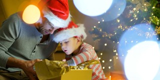 Vader en kind in kersthoeden zitten samen op de grond en openen vol enthousiasme een cadeautje, omgeven door kerstversieringen en sfeerverlichting.