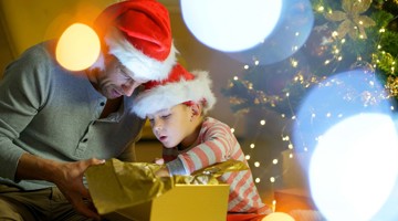 Vader en kind in kersthoeden zitten samen op de grond en openen vol enthousiasme een cadeautje, omgeven door kerstversieringen en sfeerverlichting.