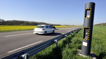 Een snelheidsmeetpaal langs een verharde weg met een passerende auto en een grasrand.
