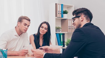 Een gesprek tussen drie mensen aan een vergadertafel, waarbij de focus ligt op een persoon die iets uitlegt terwijl de anderen luisteren en notities bekijken. Op de achtergrond staan een kantoorkast en planten.