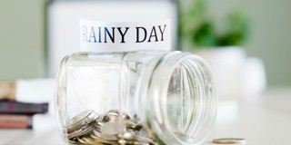 Glazen spaarpot met het etiket RAINY DAY en munten erin, op een bureau met een groene plant op de achtergrond.