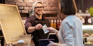 Een barista staat achter een houten toonbank en biedt een betaalterminal aan terwijl een klant een bankpas overhandigt. Achter hen zijn glazen potten met ingrediënten en flessen in verschillende kleuren te zien.