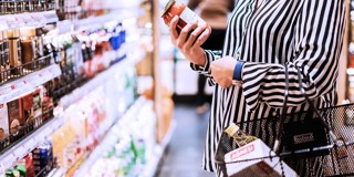 Een persoon in een gestreepte blouse houdt een pot tomatensaus vast en kijkt naar producten in een supermarkt, met een boodschappentas aan de arm.