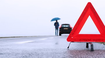 Een rode waarschuwingsdriehoek staat langs een natte weg, terwijl een persoon met een blauwe paraplu naast een stilstaande auto staat.