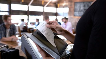 Een hand houdt een bonnetje boven een zwart informatiepaneel in een druk restaurant met mensen op de achtergrond.