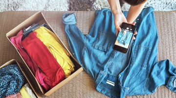 Une personne prend en photo une chemise en denim avec un smartphone, tandis qu'une boîte contenant des vêtements colorés est posée à côté d'eux sur un lit.