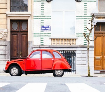 Een felrode vintage auto naast een historische gevel met groene tegels en houten deuren.