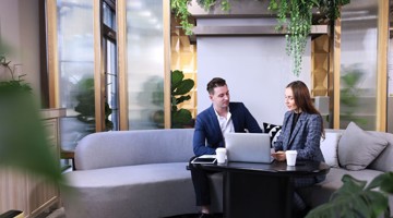 Deux personnes en tenue professionnelle sont assises à une table avec des ordinateurs portables et des tasses de café, entourées de plantes dans un espace de bureau moderne.
