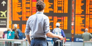 Un homme avec une valise regarde un tableau d'informations à une gare animée, affichant des horaires de départ en orange vif.
