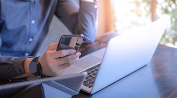 Personne tenant un smartphone tout en regardant un ordinateur portable sur un bureau, manifestant de l'inquiétude.