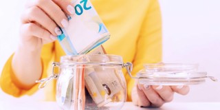 Een hand plaatst eurobankbiljetten in een glazen pot vol met geld.