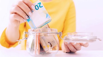 Een hand plaatst eurobankbiljetten in een glazen pot vol met geld.