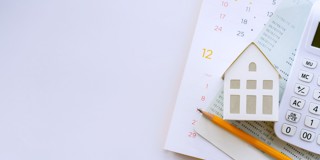 Un calendrier blanc avec une petite maison en bois, une calculatrice et un crayon, sur un fond clair.