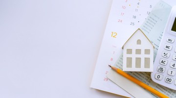 Un calendrier blanc avec une petite maison en bois, une calculatrice et un crayon, sur un fond clair.