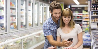 Un homme et une femme se tiennent près l'un de l'autre dans un supermarché, tandis que l'homme regarde un smartphone et la femme jette un œil par-dessus son épaule. Derrière eux, on peut voir des congélateurs et des étagères avec des produits.